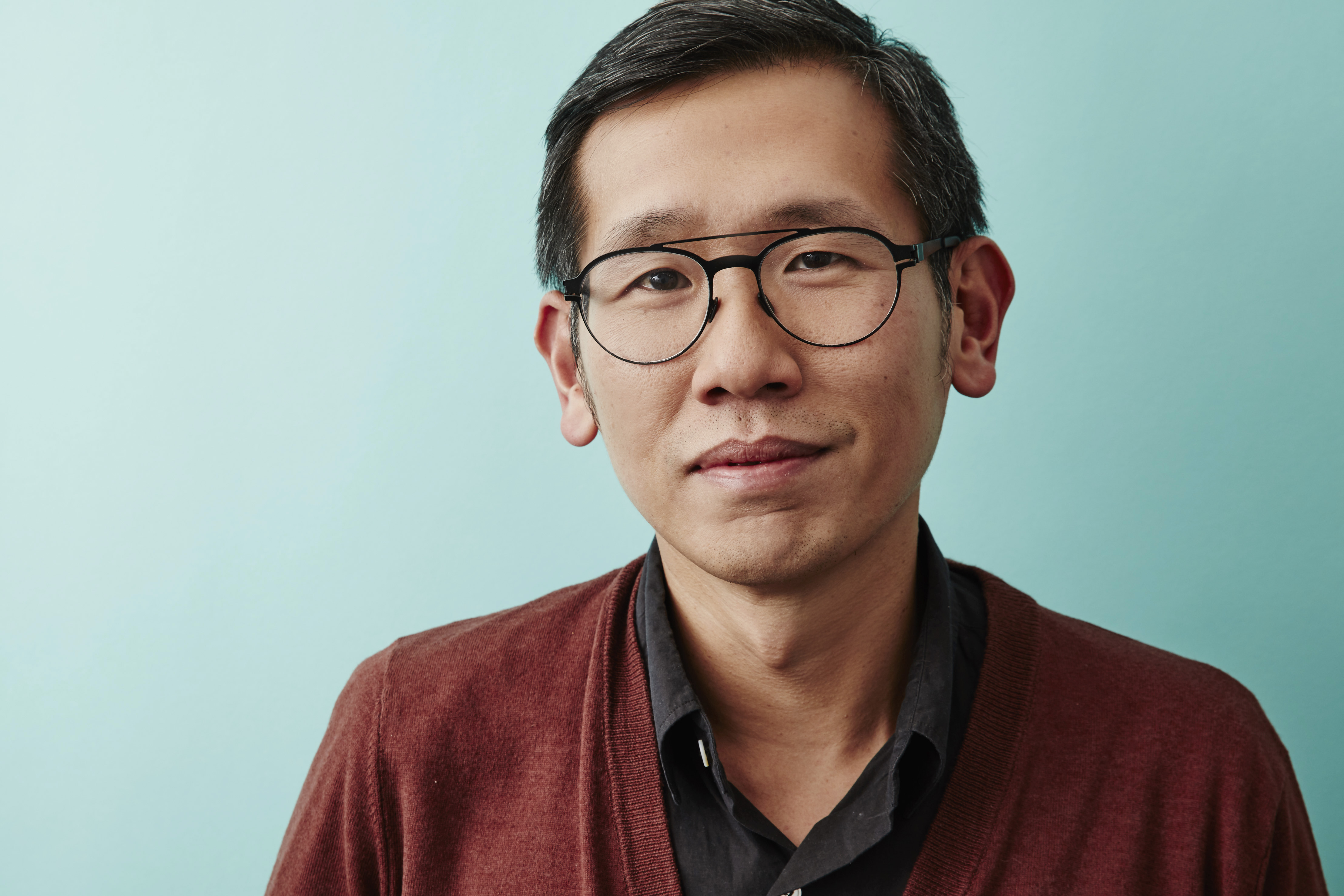 A photo of Dennis Lim, an Asian man wearing glasses and a red sweater, smiling with his mouth closed. Behind him is a robin's egg blue backdrop.