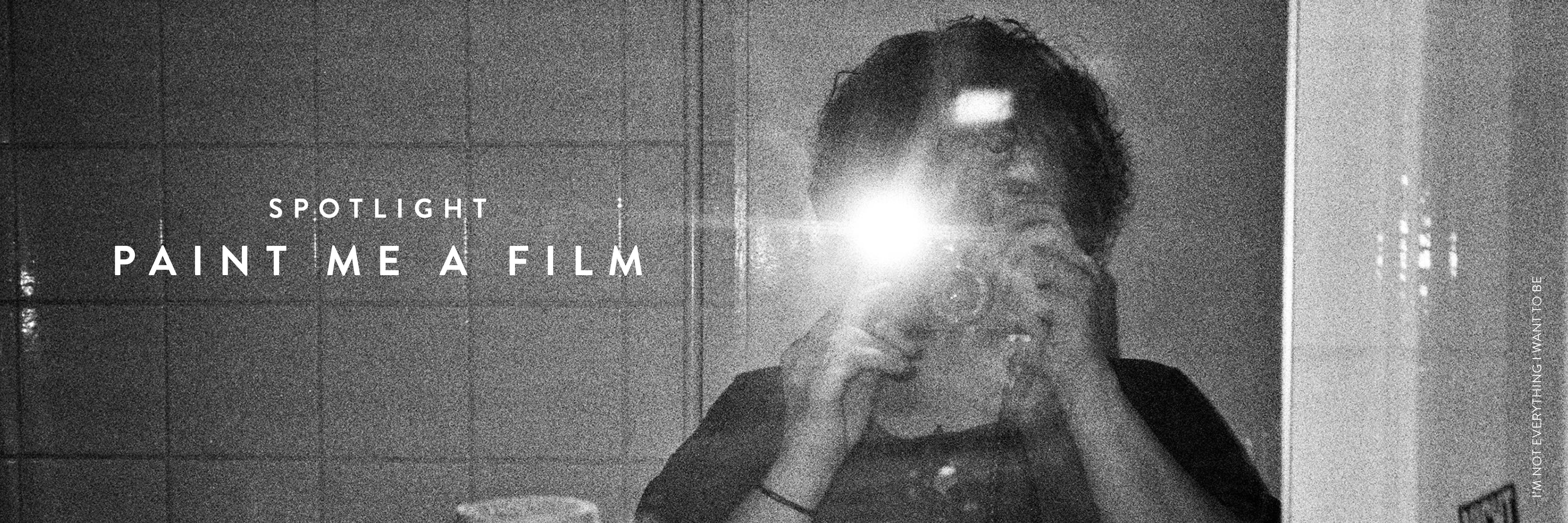 Black and white photograph of a woman taking a photo in the mirror, the camera flash obscuring her face. White text on the left of the image reads: Spotlight PAINT ME A FILM