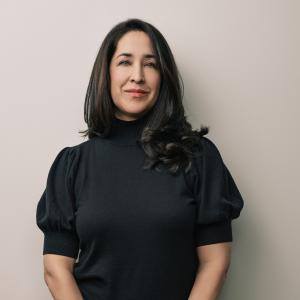 Michelle Chawla, a woman with long dark hair wearing a black blouse, stands against a taupe background and smiles at the camera with her mouth closed.